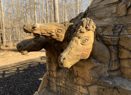 Sculpture of two horse heads emerging from a rocky surface, surrounded by bare trees in a park setting.