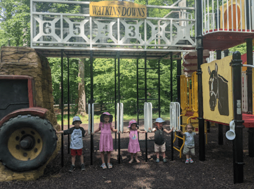 Five children play together at a colorful playground, smiling and holding onto a structure in a green park.