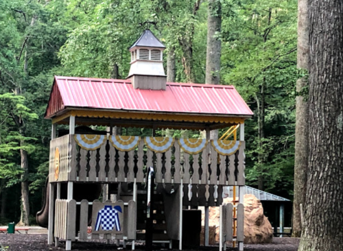 A wooden playhouse with a red roof, surrounded by trees, featuring a slide and climbing structure.