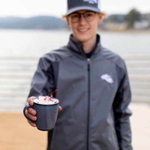 A person in a gray jacket holds a mug topped with whipped cream and sprinkles, with a scenic background.