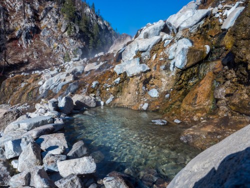 A clear, natural hot spring surrounded by rocky terrain and mountains under a bright blue sky.