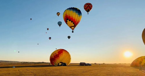 Colorful hot air balloons ascend into a clear sky at sunrise, with a golden landscape below.