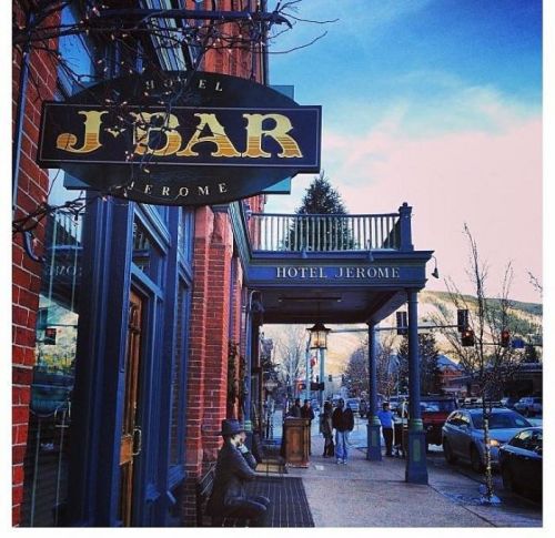 A street view of Hotel Jerome with a sign for J-Bar, featuring people walking and a snowy mountain backdrop.