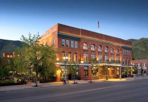 Historic brick building with large windows, trees, and bicycles outside, set against a mountain backdrop at dusk.