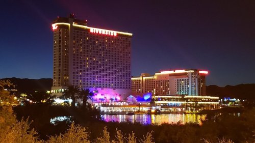 A brightly lit hotel at night, reflecting on a calm body of water, surrounded by mountains and palm trees.