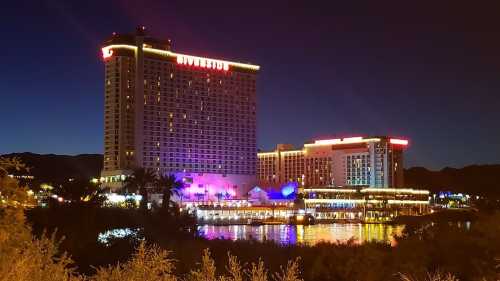 A brightly lit hotel at night, reflecting on a calm body of water, surrounded by mountains and palm trees.