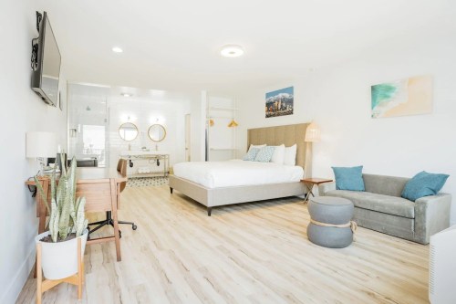 Bright, modern hotel room featuring a queen bed, desk, sofa, and kitchenette with stylish decor and natural light.