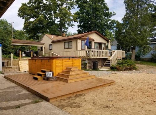 A cozy house with a wooden deck and hot tub, surrounded by trees and sandy ground.