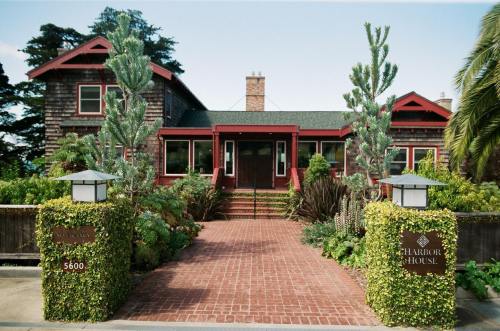 A charming wooden house with a green roof, surrounded by lush plants and a brick pathway leading to the entrance.