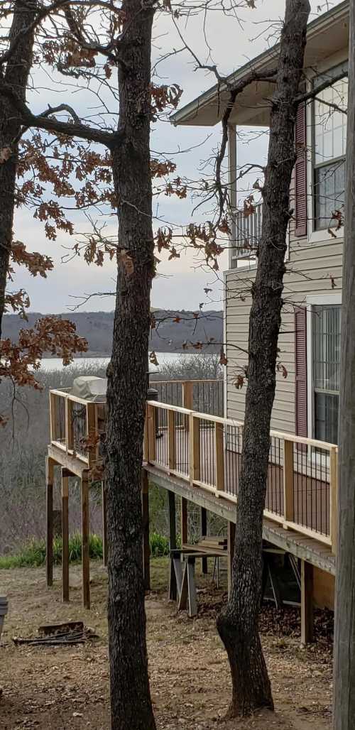 A wooden deck extends from a house, surrounded by trees, overlooking a lake in the distance under a cloudy sky.