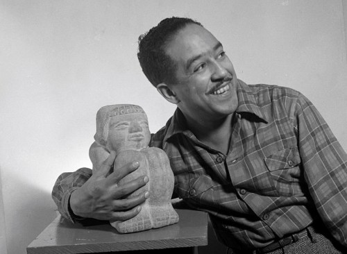 A smiling man in a plaid shirt poses with a stone sculpture of a seated figure.