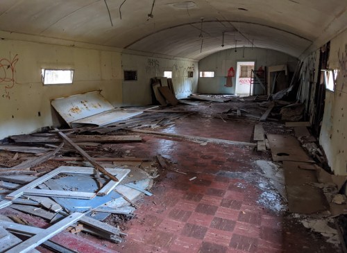 An abandoned, dilapidated room with debris, broken flooring, and graffiti on the walls. Natural light enters through a window.
