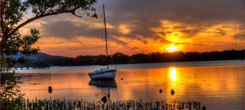 A serene sunset over a calm lake, with a sailboat anchored and reflections shimmering on the water.
