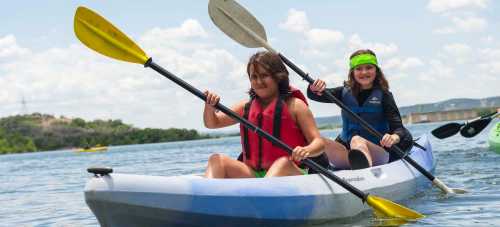 Two children paddle a kayak on a sunny day, wearing life jackets and enjoying the water.