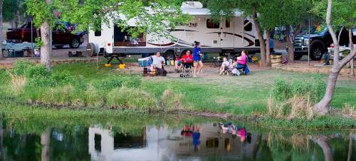 A family gathers by a lake near their RV, enjoying a camping trip with chairs and a fire pit. Reflections in the water.