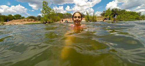 A girl smiles while swimming in a lake, surrounded by greenery and people enjoying the sunny day.