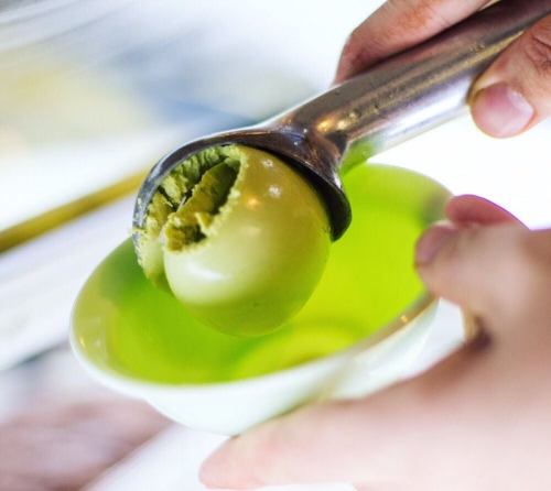 A hand holding a metal ice cream scoop with a ball of green ice cream above a small green bowl.