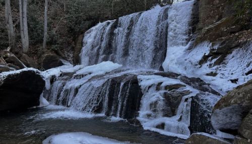 A frozen waterfall cascading over rocky ledges, surrounded by trees in a serene natural setting.
