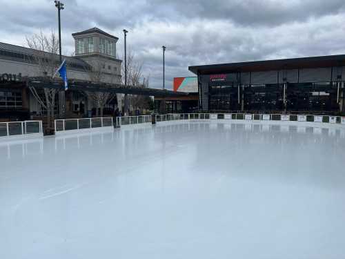 An empty outdoor ice skating rink under a cloudy sky, surrounded by buildings and trees.