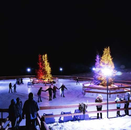 A nighttime ice skating rink with colorful trees and people skating, surrounded by festive lights.