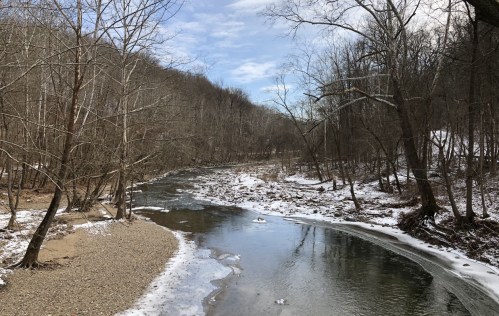 A serene river flows through a winter landscape with bare trees and patches of snow along the banks.
