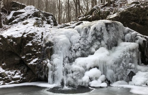 A frozen waterfall cascading over rocky terrain, surrounded by snow-covered trees in a winter landscape.