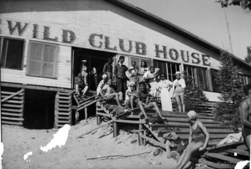 A group of people in swimsuits poses on the steps of a building labeled "Wild Club House" by a sandy area.
