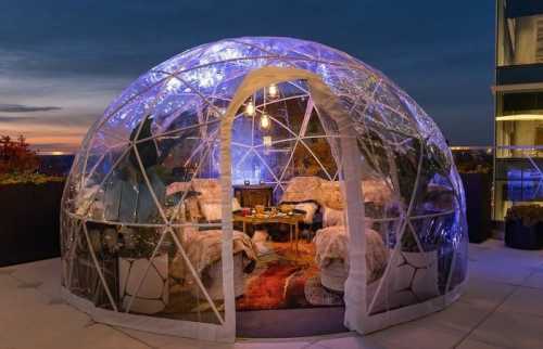 A cozy, illuminated igloo-style dome on a rooftop, featuring plush seating and a warm ambiance at dusk.