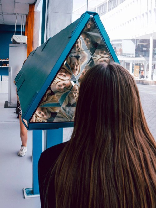 A person with long hair looks into a triangular mirror, reflecting multiple images of their face.