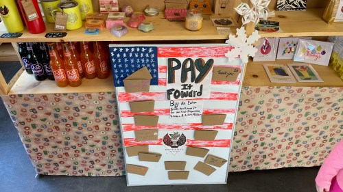 A display board with a U.S. flag design, promoting a "Pay It Forward" initiative with envelopes for donations.