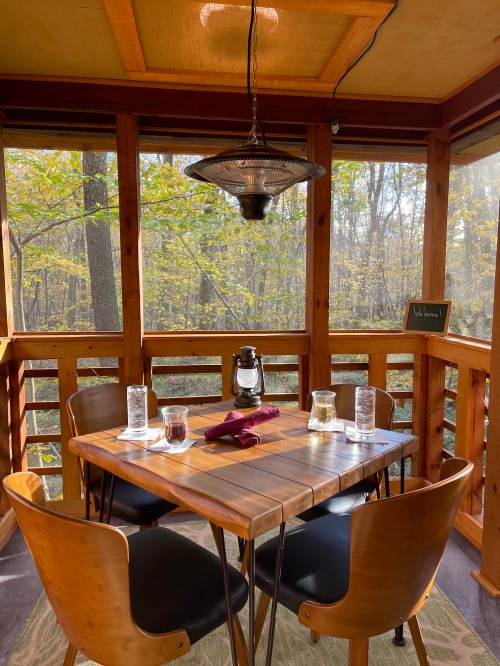 A cozy dining area with a wooden table, chairs, and lantern, surrounded by large windows showcasing autumn foliage.