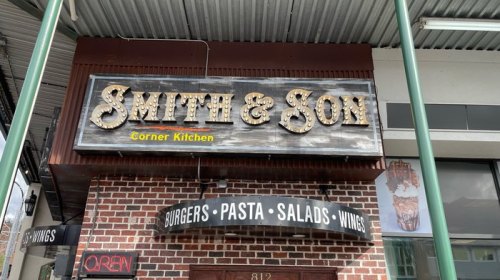 Sign for "Smith & Son Corner Kitchen" featuring a brick facade and a menu of burgers, pasta, salads, and wings.