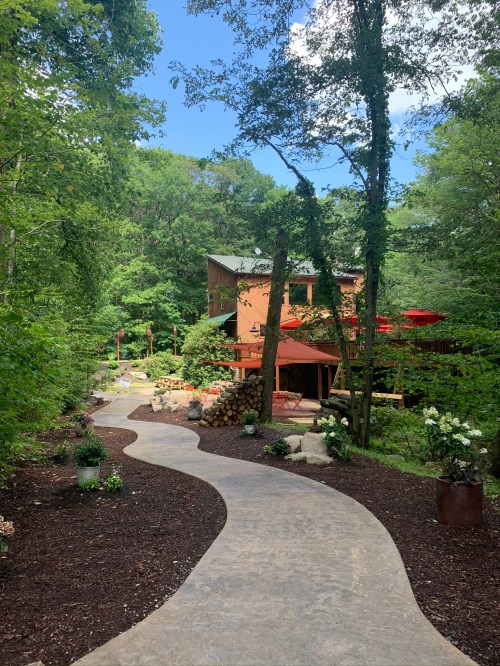 A winding stone path leads through lush greenery to a cozy house surrounded by trees and garden features.