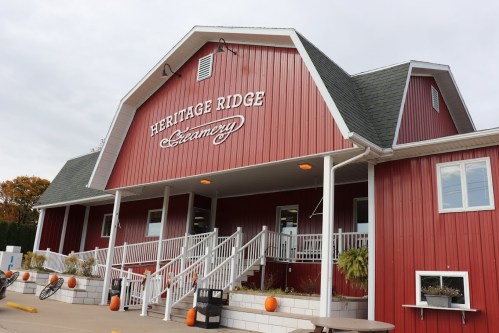 Red barn-style building with "Heritage Ridge Creamery" sign, white railings, and pumpkins on the porch.