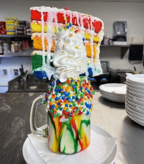 A colorful milkshake topped with whipped cream and a slice of rainbow cake, adorned with sprinkles.