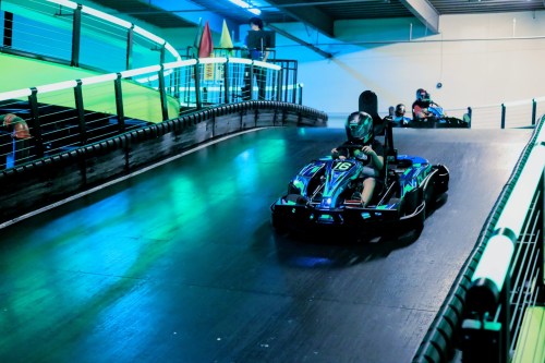 A go-kart racer navigates a curved track in a brightly lit indoor racing facility.