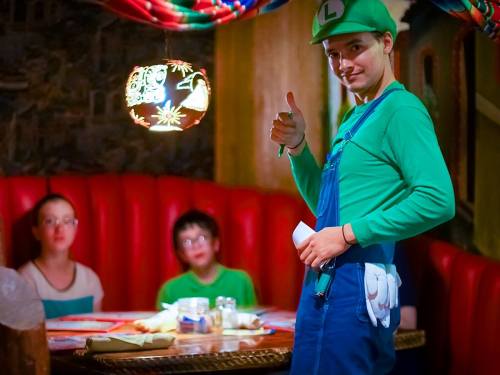 A person dressed as Luigi gives a thumbs up in a restaurant, with two children seated at a table in the background.