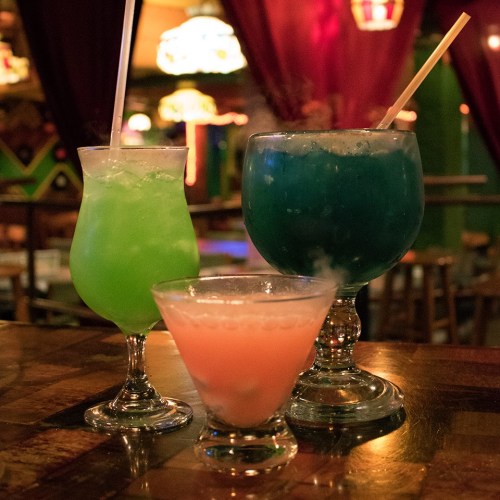 Three colorful cocktails on a wooden table: a green drink, a blue drink, and a pink drink, all with straws.