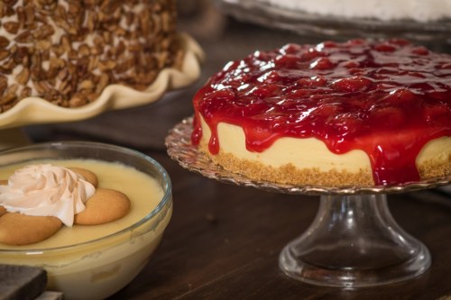 A cheesecake topped with cherry glaze next to a bowl of creamy dessert with cookies and whipped cream.