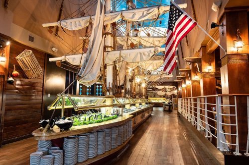A nautical-themed restaurant interior featuring a ship model, buffet area, and American flag. Warm wooden decor.