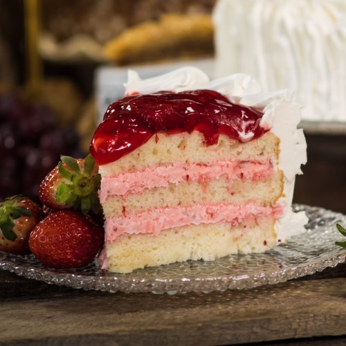 A slice of layered cake with pink frosting and cherry topping, garnished with fresh strawberries on a glass plate.