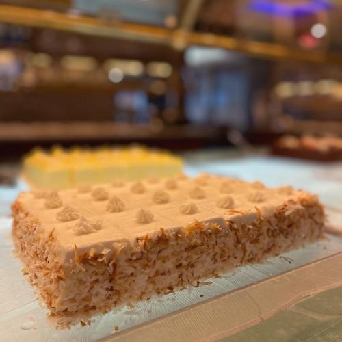 A close-up of a rectangular cake topped with cream and sprinkled with toasted coconut, displayed in a bakery.
