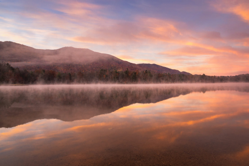 Serene lake at sunrise, reflecting mountains and colorful sky, with mist rising from the water's surface.