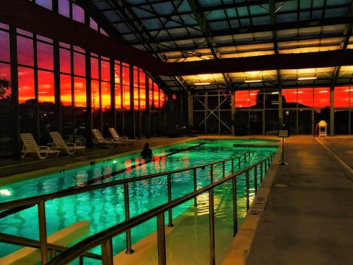 Indoor pool with turquoise water, surrounded by lounge chairs, under a vibrant sunset sky with orange and purple hues.