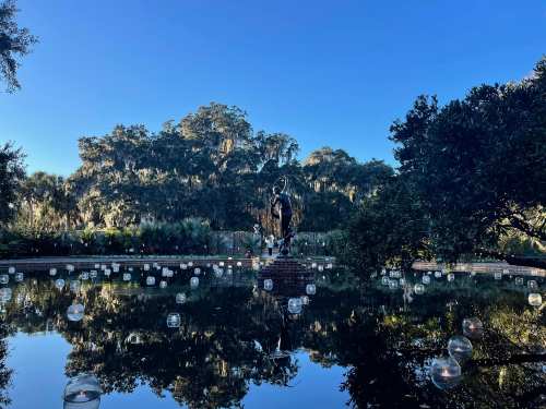A serene pond reflects a statue surrounded by trees and floating lanterns under a clear blue sky.
