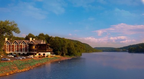 Scenic riverside view featuring a lodge, trees, and a calm waterway under a blue sky with clouds.