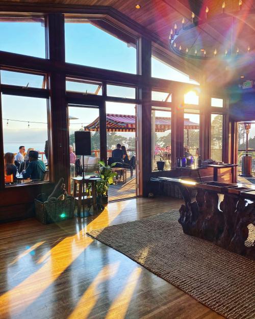 A sunlit interior of a cozy venue with large windows, showcasing a scenic view and warm wooden decor.
