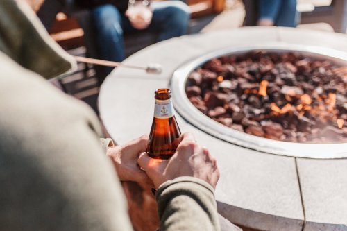 A person holds a beer bottle while sitting near a fire pit with glowing embers.
