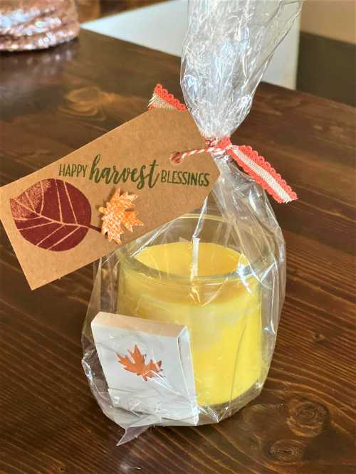 A wrapped candle with a tag that reads "Happy Harvest Blessings," featuring autumn leaves and a decorative ribbon.