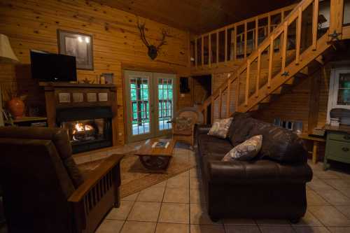 Cozy cabin interior with wooden walls, a fireplace, a sofa, and a staircase leading to an upper level.